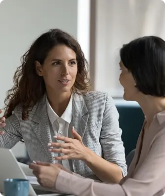 Two women engaged in conversation at a desk in a modern office environment.